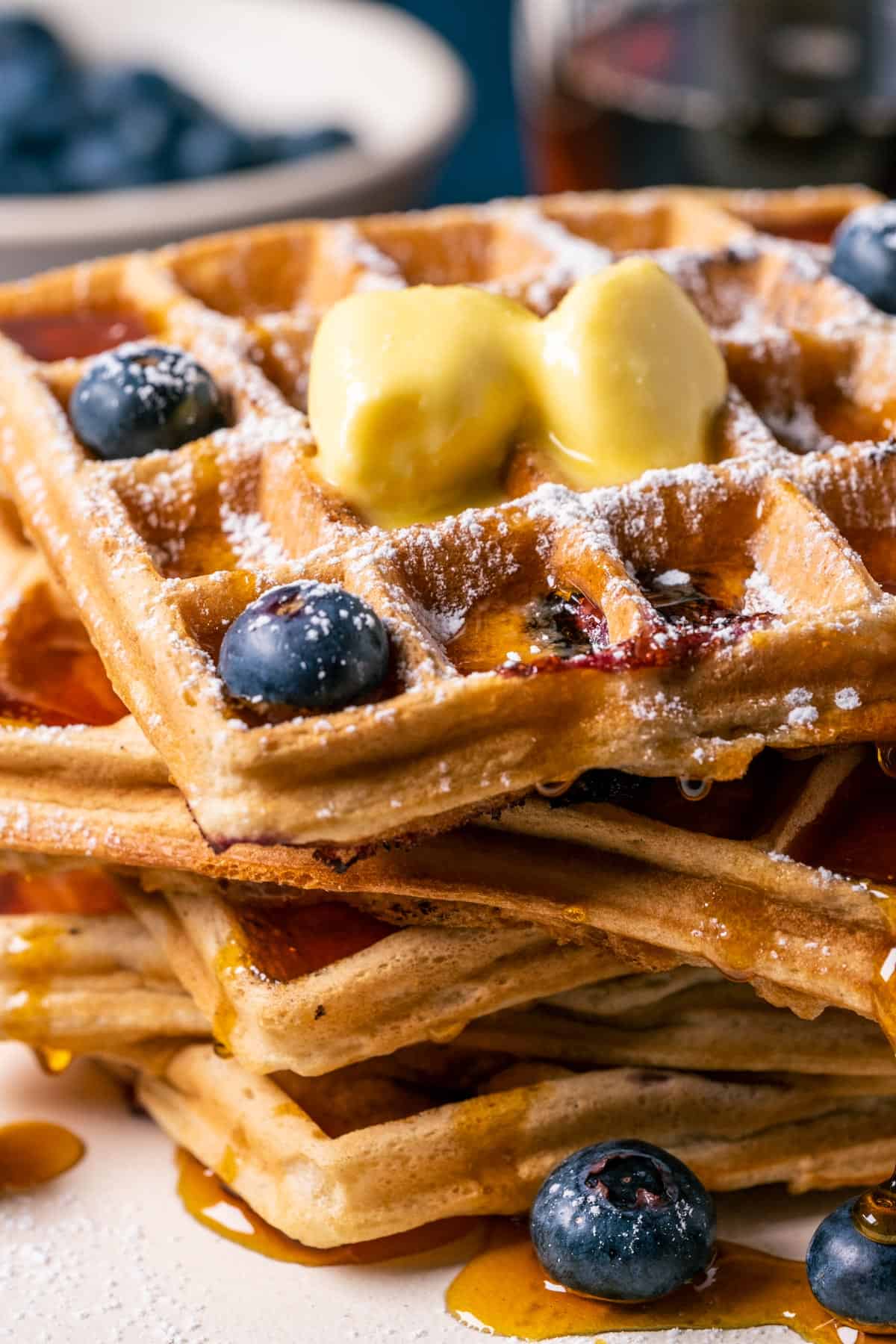 Stack of blueberry waffles topped with vegan butter, powdered sugar and syrup on a white plate.