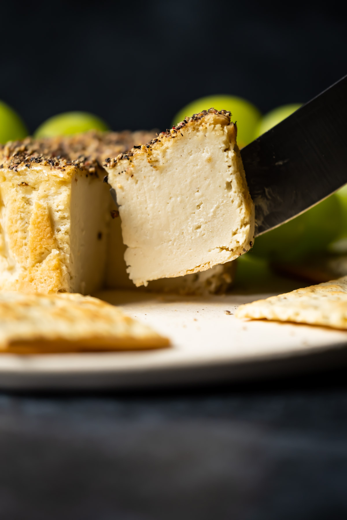 Slice of vegan camembert ready to serve from a white plate with crackers and grapes.