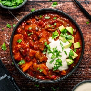 Vegan chili with cashew cream, chopped cilantro and diced avocado in a black bowl.