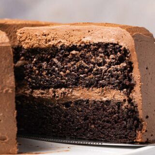 Vegan chocolate cake on a white cake stand with one slice cut and ready to serve.