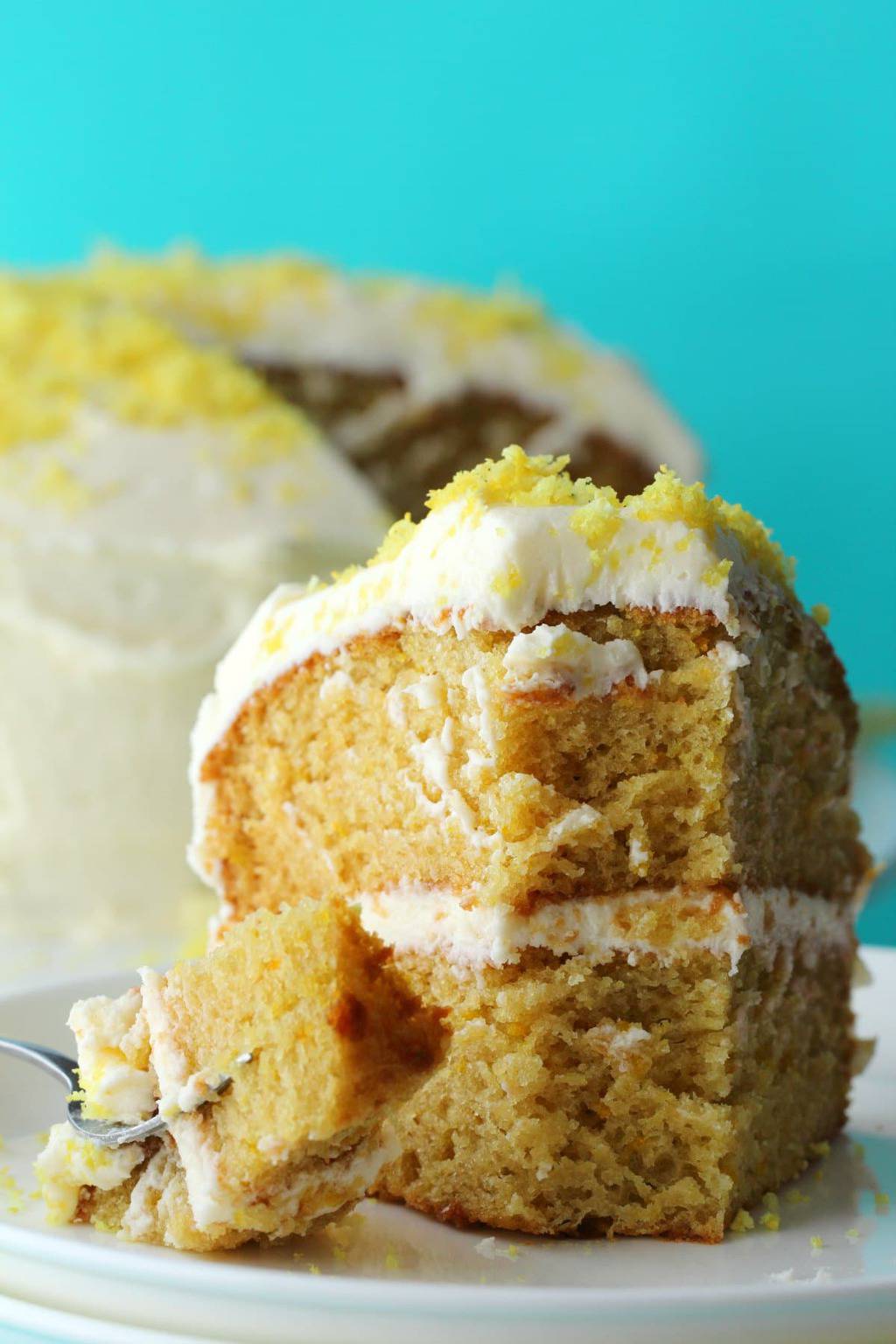 A slice of vegan lemon cake on a white plate with a forkful of cake on a cake fork next to it. 