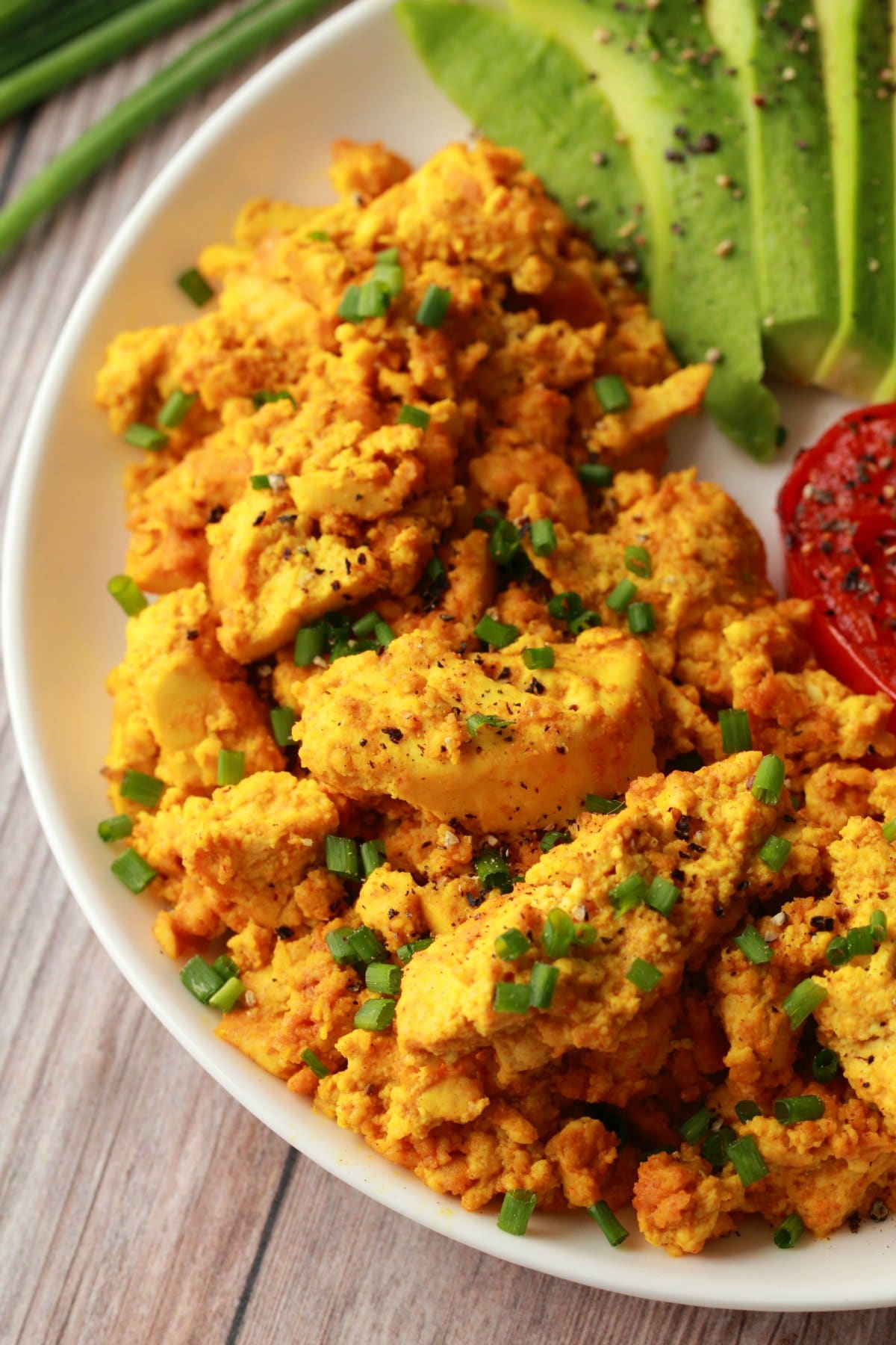 Tofu scramble topped with chopped chives on a white plate with fried tomato and sliced avocado. 