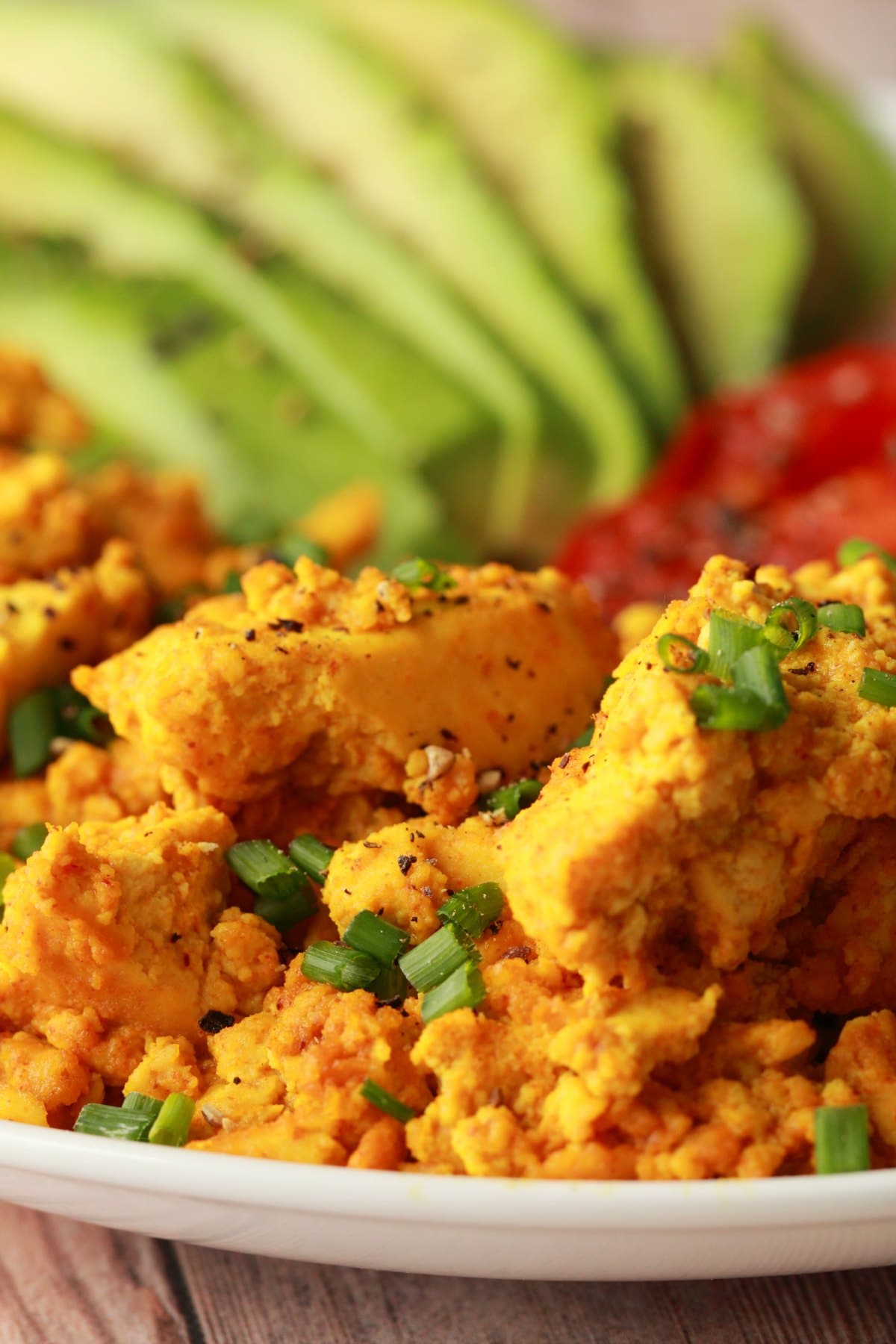 Tofu scramble topped with chopped chives on a white plate with sliced avocado and fried tomato. 