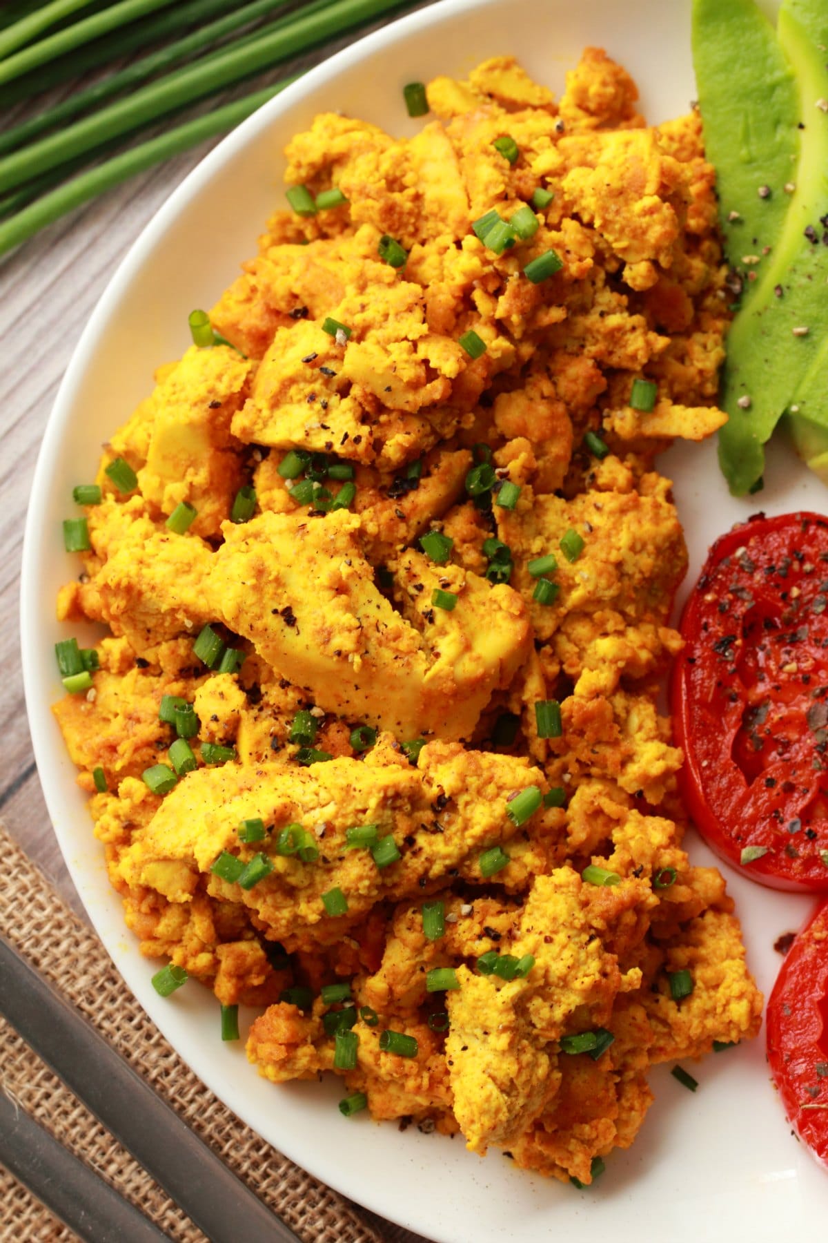 Tofu scramble topped with chopped chives on a white plate with fried tomato and sliced avocado. 